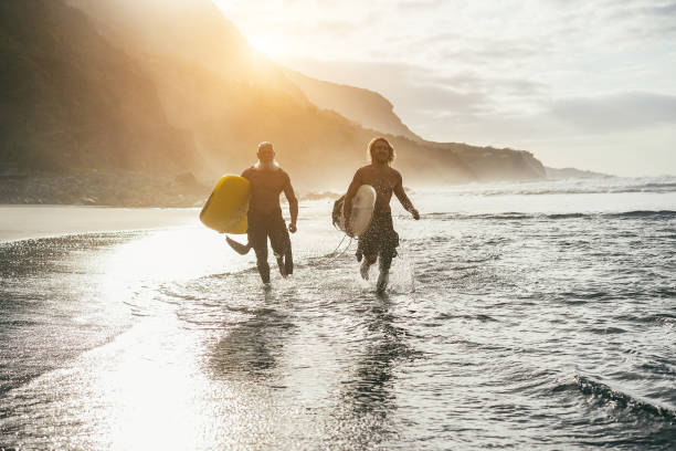 amigos surfistas multigeracionais se divertindo correndo na praia após a sessão de surf - foco suave nas pranchas de surf - surfing beach family father - fotografias e filmes do acervo