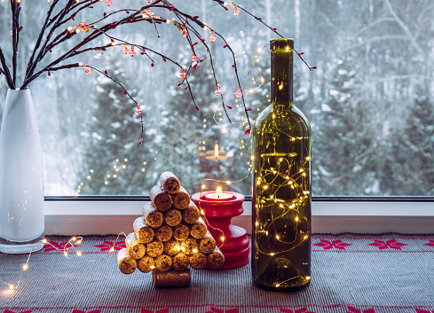 Christmas decoration set with wine bottle filled with micro led party lights and spruce tree made with used wine corks, behind is window with snowy countryside forest.