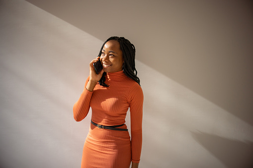 Portrait of modern woman of Black ethnicity, in orange dress, talking on mobile phone, on white background