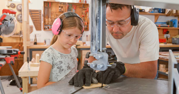 Daughter and her father cutting wooden plank with electric saw while wearing safety glasses in carpentry workshop.