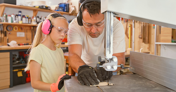 Daughter and father cutting wooden plank with electric saw while wearing safety glasses in carpentry workshop.