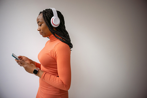 Side view of young woman of Black ethnicity, in orange dress, with wireless headphones on, holding mobile phone, while choosing song on her playlist