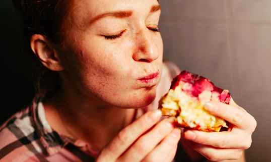 Portrait of a red-haired woman in her kitchen. She afforded herself a slice of raspberry pie. She eats the cake with her hands. She enjoys this moment.