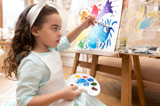 Little girl in white apron drawing on a drawing easel.