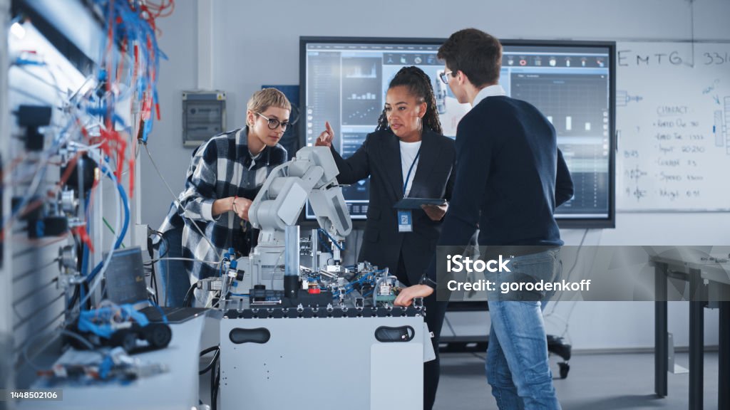 In Robotics Development Laboratory: Black Female Teacher and Two Students Work With Prototype of Robotic Hand. Young Student Telling Something with Smile and Brainstorming with Her Team. Engineer Stock Photo