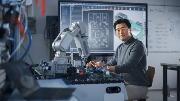 istock Asian Male Engineer Sitting at Table And Looking at Camera While Woking on New Engineering Project. Robot is Moving Under his Control. Education and Robotics In Modern University Concept. 1448503128
