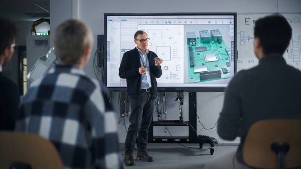 istock Diverse Team of Computer Design Students Talking at University with Teacher, Watching Interactive TV that shows 3D Concept Model of Printed Circuit Board. Education Concept 1448503158