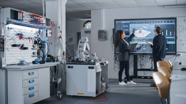 istock Female Black Developer Looking at Big Screen and Telling her Ideas to Male Colleague. Robotic Arm Moving in Background. Science and Innovative Technologies on Modern Factory Concept. 1448503503