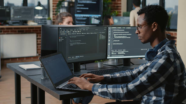 istock Portrait of tired programer rubbing eyes while typing on laptop sitting at desk with multiple screens parsing code 1448505771
