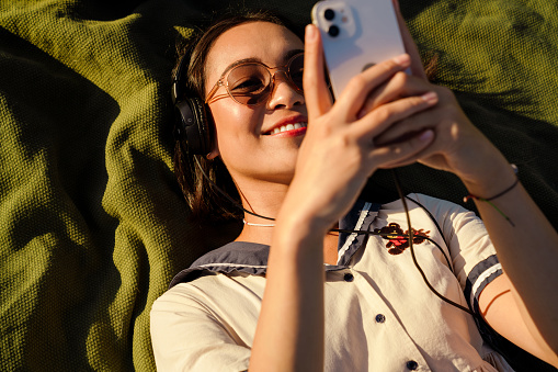 Top view of joyful young asian girl listening music with headphones and mobile phone while laying on blanket outdoors