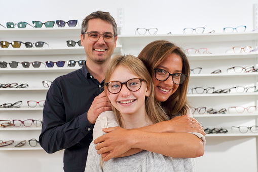 Father helping his daughter to buy new eyeglasses. Playful and smiling family in optics shop. Looking at some frames.