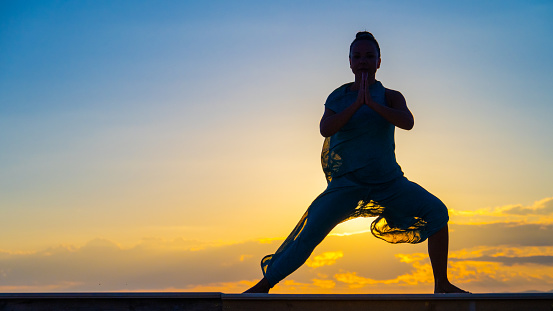 Silhouette of woman practising goddess yoga on deck during sunset.