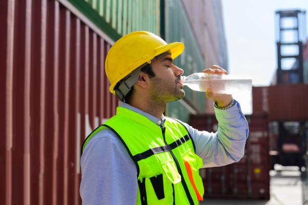 istock Industrial worker in safety hard hat and vest drinking clear water with containers stack background 1448528833