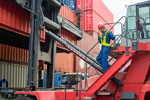 Engineer technician walking down on forklift container using walkie-talkie to communicate and control loading containers with logistics team in import and export zone at cargo containers yard