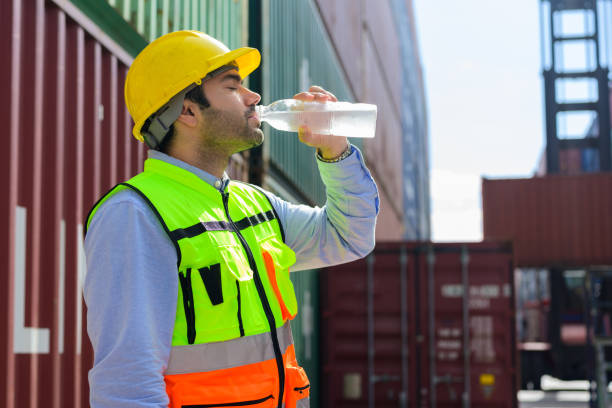 istock Industrial worker in safety hard hat and vest drinking clear water with containers stack background 1448530773