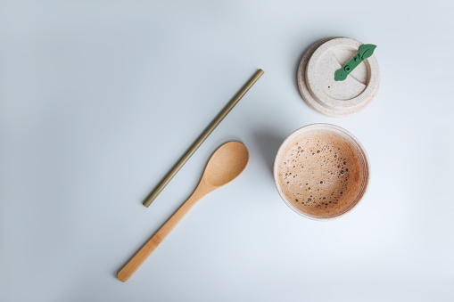 Coffee with foam in an eco glass on a white table with a bamboo straw and wooden spoon. Flat lay