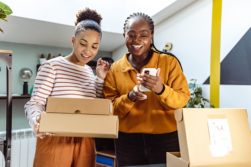 Diverse female friends just received their online orders. They are curious to open the package. Young African-American woman using smart phone for checking online orders