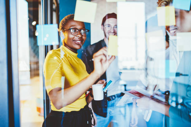 istock Group of happy hipster guys making notes on colorful memo sticks at glass wall while creating interesting ideas, cheerful multiracial male and female smiling while working with startup project 1448535570