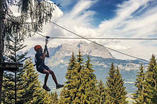 Teenage boy enjoying high ropes course in adventure park. \nThe boy is wearing safety harness and a helmet.\nThe boy is zip lining from one platform to another. Spectacular view of Austrian Alps mountains in the background.\nSunny summer day.\nCanon R5