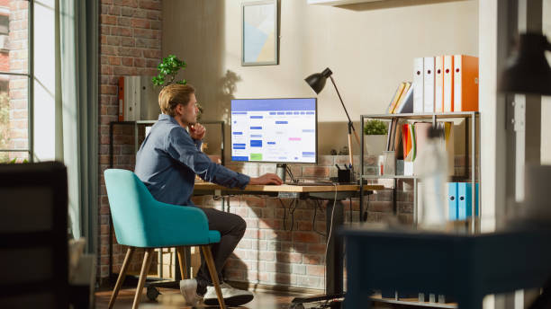 istock Remote Work Concept: Man Using Desktop Computer With Office Software to Check Schedule in Calendar. Young Male Planning Business Meetings in Stylish Loft Apartment with Big Window. 1448542135