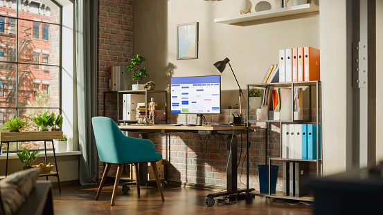 Desktop Computer Monitor Standing on Height Adjustable Desk with Office Software on Display. Cozy Empty Loft Apartment with a Lamp, Notebooks and Smartphone on the Table. Home Office Concept.