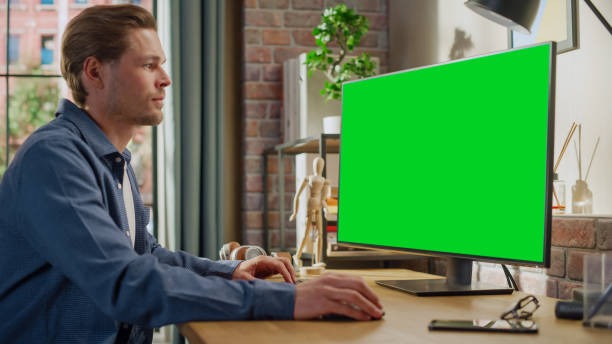 istock Young Handsome Man Working from Home on Desktop Computer with Green Screen Mock Up Display. Male Checking Corporate Accounts, Messaging Colleagues. Loft Living Room with Big Window. 1448542573