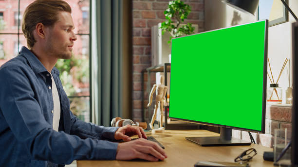 istock Young Handsome Man Working from Home on Desktop Computer with Green Screen Mock Up Display. Male Checking Corporate Accounts, Messaging Colleagues. Loft Living Room with Big Window. 1448542623