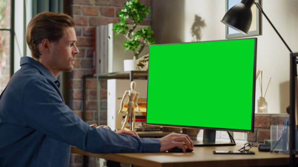 istock Young Handsome Man Working from Home on Desktop Computer with Green Screen Mock Up Display. Male Checking Corporate Accounts, Messaging Colleagues. Loft Living Room with Big Window. 1448542661
