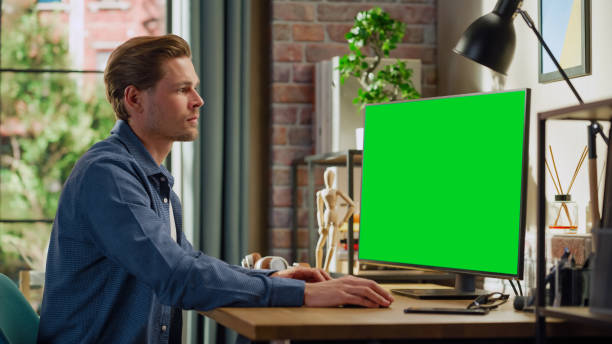 istock Young Handsome Man Working from Home on Desktop Computer with Green Screen Mock Up Display. Male Checking Corporate Accounts, Messaging Colleagues. Loft Living Room with Big Window. 1448542699