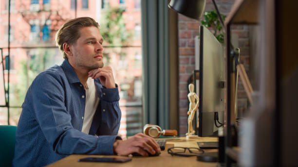 istock Portrait of a Young Handsome Man Sitting at Home, Working on Desktop Computer. Creative Male Checking Social Media, Browsing Internet. Living Room in Bright Loft Apartment. 1448542777