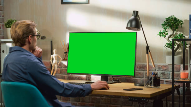 istock Young Handsome Man Working from Home on Desktop Computer with Green Screen Mock Up Display. Creative Male Checking Social Media, Browsing Internet. Living Room in Bright Loft Apartment. 1448542918