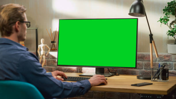 istock Young Handsome Man Working from Home on Desktop Computer with Green Screen Mock Up Display. Creative Male Checking Social Media, Browsing Internet. Living Room in Bright Loft Apartment. 1448542989