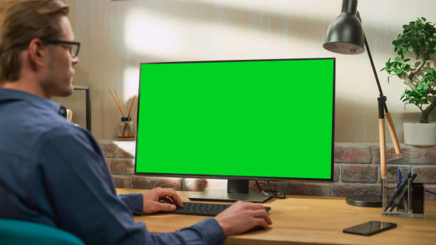 istock Young Handsome Man Working from Home on Desktop Computer with Green Screen Mock Up Display. Creative Male Checking Social Media, Browsing Internet. Living Room in Bright Loft Apartment. 1448543600