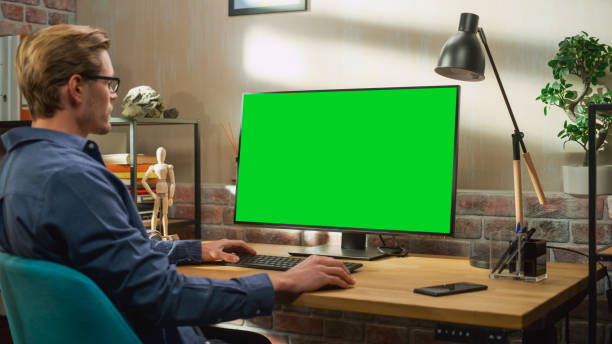 istock Young Handsome Man Working from Home on Desktop Computer with Green Screen Mock Up Display. Creative Male Checking Social Media, Browsing Internet. Living Room in Bright Loft Apartment. 1448543628