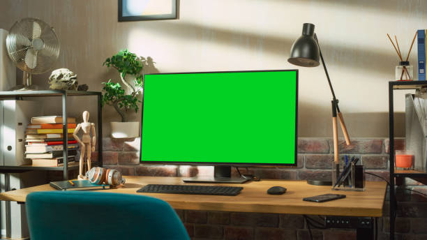 istock Desktop Computer Monitor Standing on a Desk with a Green Screen Chromakey Mock Up Display. Cozy Empty Loft Apartment with a Lamp, Notebooks, Smartphone and Headphones on the Table. 1448543662