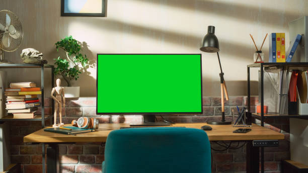 istock Desktop Computer Monitor Standing on a Table with a Green Screen Chromakey Mock Up Display. Cozy Empty Loft Apartment with a Lamp, Notebooks, Smartphone and Headphones on the Table. 1448543717