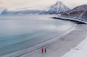 Aerial image of woman and man walking at the scenic turqouse beach in snow with mountain view in Norway