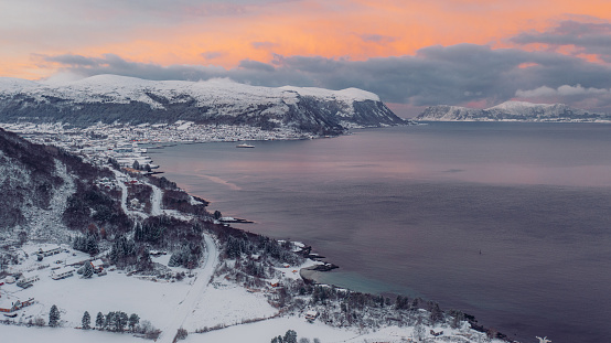 Drone high-angle photo of the town by the crystal blue ocean water and view of snowcapped mountains during dramatic pink december sunset in More og Romsdal, Scandinavia