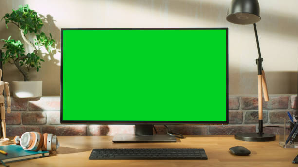 istock Desktop Personal Computer Monitor Standing on a Table with a Green Screen Chromakey Mock Up Display. Cozy Empty Loft Apartment with a Lamp, Notebooks and Headphones on the Table. 1448543876