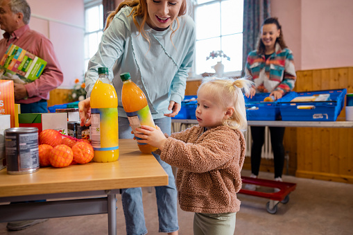 Medium shot of a mother and daughter picking up food together at a food bank. It is being run by volunteers at a community church in the North East of England. The focus is on a young girl is holding a bottle of juice.