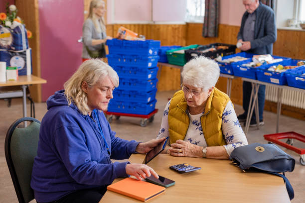 Meeting with a Financial Advisor Two older women at a community church where a food bank is being run by volunteers in the North East of England. They are sitting at a table together, using a calculator to work out budgets, helping each other to save money during the inflation. community center food stock pictures, royalty-free photos & images