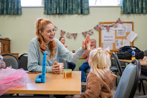 Mother and her young daughter relaxing at a community church in the North East of England which is functioning as a warm hub. People in need are warming up there due to heating prices/inflation. The young girl is watching a movie on a digital tablet while her mother drinks a cup of tea. They are high-five.