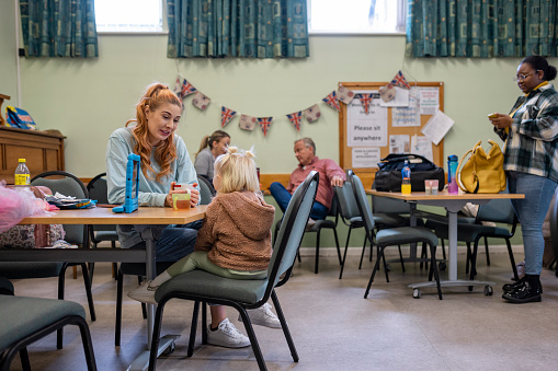 Wide shot of a mother and her young daughter relaxing at a community church in the North East of England which is functioning as a warm hub. People in need are warming up there due to heating prices/inflation. The young girl is watching a movie on a digital tablet while her mother drinks a cup of tea. Other people are also using the space behind them.