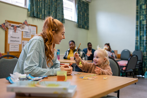 Mother and her young daughter relaxing at a community church in the North East of England which is functioning as a warm hub. People in need are warming up there due to heating prices/inflation. The young girl is playing a fishing game with a miniature rod, having fun. Other people are out of focus behind them.