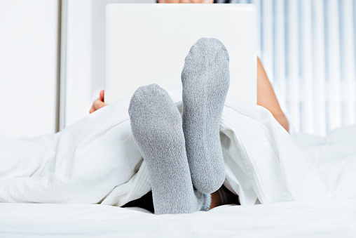 Close up of caucasian persons feet wearing gray socks in bed with white sheet using laptop.