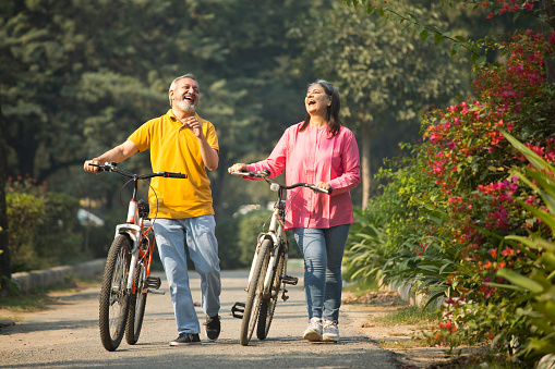 Carefree senior couple talking and laughing while walking with bicycles on road amidst plants in park during weekend