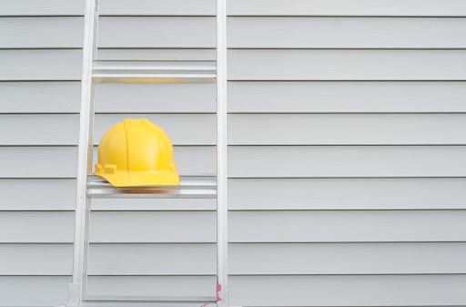 Yellow hardhat on ladder with house siding in background.