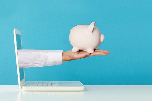 Hand holding white piggy bank  is reaching out of white laptop screen on white table in front of blue wall. Representing online personal finance consultancy
