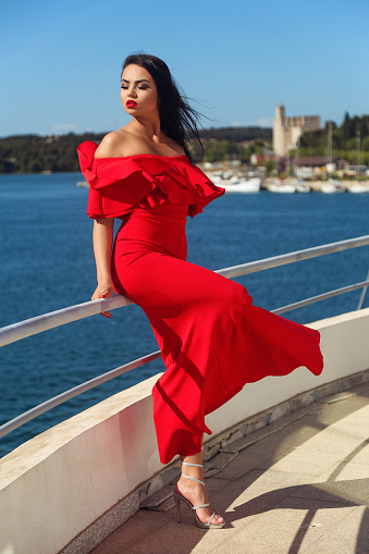 Beautiful young black-haired woman in a long red dress is enjoying herself on a yacht looking at the sea