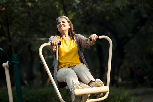 Full length of smiling Indian senior woman looking away while sitting on exercise equipment at playground in morning
